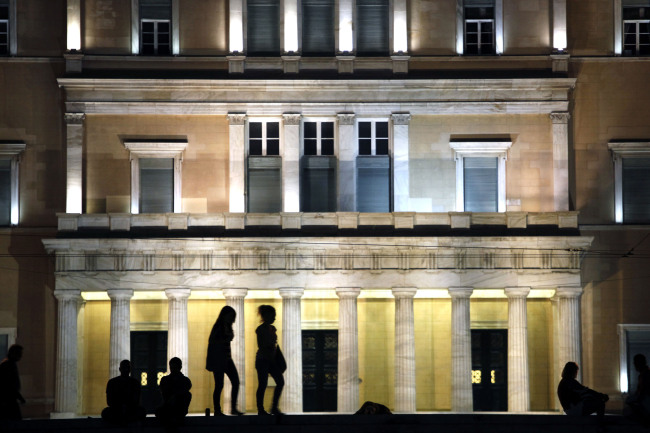 Pedestrians stand in front of the illuminated facade of the Greek parliament building in Athens.  Bloomberg