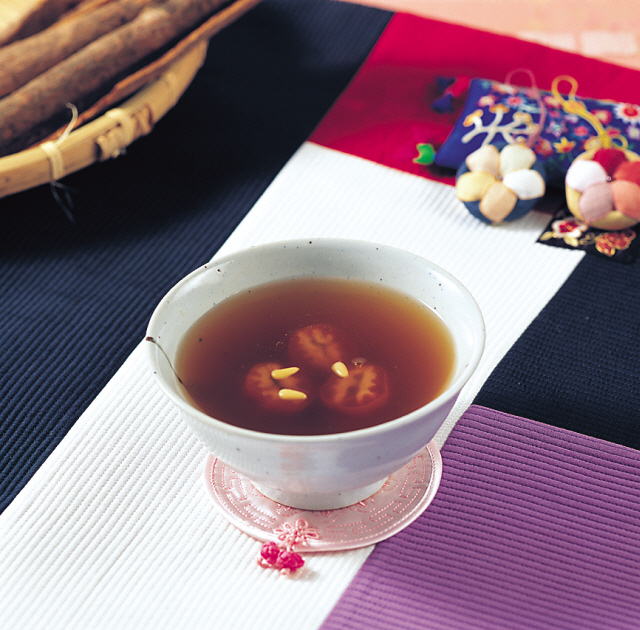 Sujeonggwa served in a small speckled ceramic tea bowl with pine nuts floating on the surface, set on a traditional Korean textile placemat with silk norigae pouches and a bamboo basket of ingredients