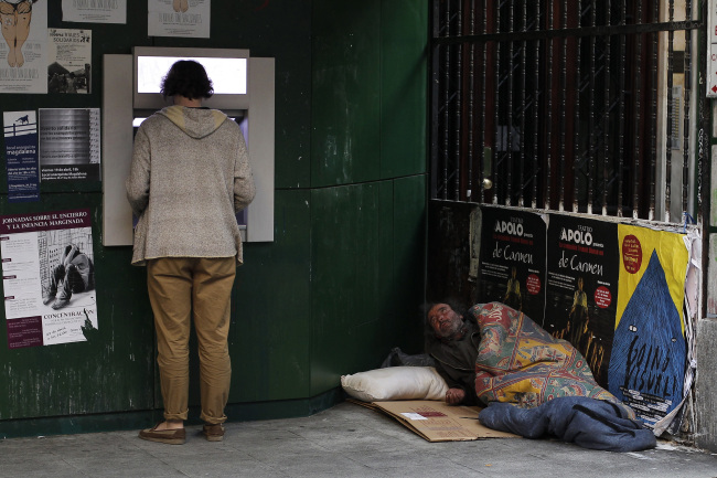 A man uses an ATM machine as a homeless man begs for alms from his makeshift bed in Madrid on Monday.   AP-Yonhap News