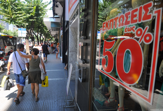 Pedestrians pass a store advertising half price discounts in its window display in Thessaloniki, Greece.     Bloomberg