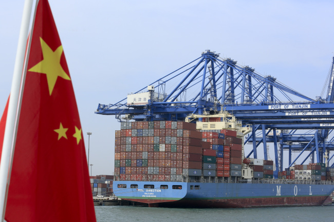 A Chinese flag flies on a vessel moving past shipping containers being unloaded at a Tianjin Port Group Co. dock in Tianjin on Thursday.   Bloomberg