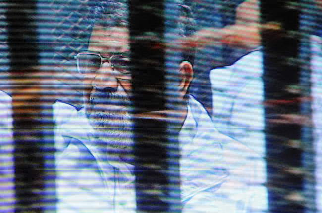 Egypt’s ousted President Mohammed Morsi, in a soundproof barred glass cage, is seen on a monitor set up outside a courtroom where he and 35 others are facing charges of conspiring with foreign groups and undermining national security in Cairo on Sunday.   AP-Yonhap
