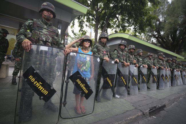 A young boy wearing a military helmet and sunglasses salutes as he poses with Thai soldiers blocking a road to prevent protesters from rallying against the military coup near the Victory Monument in Bangkok on Friday.   EPA-Yonhap
