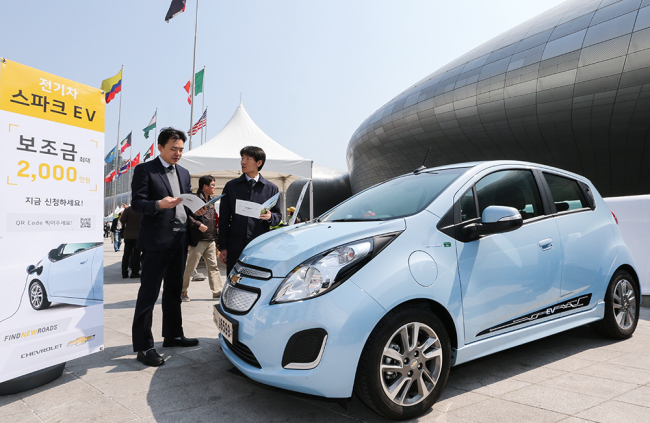 People look at a brochure for GM’s Chevrolet Spark EV displayed outside Dongdaemun Design Plaza in Seoul. Chevrolet is subsidizing the price of the Spark EV to 2.19 million won ($20,000) at its sales outlets in Seoul until June 5.   GM Korea
