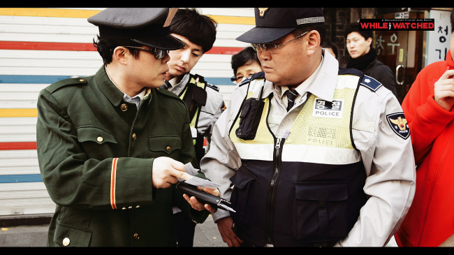 Justice for North Korea protestors are quizzed by police in a still from “While they Watched.”   Square Eyed Pictures