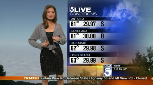 A screen capture of a KTLA-TV weather forecast where weather anchor Liberte Chan puts on a gray sweater handed over by cohost Chirs Burrous from off camera. (KTLA)