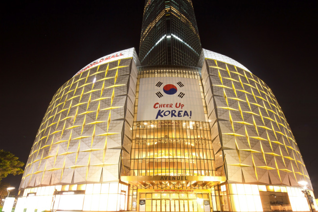 금빛 조명을 밝힌 롯데월드타워와 에비뉴엘 파사드에 부착된 대형 태극기와 응원 메시지.