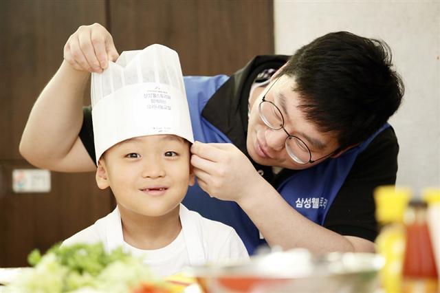 [최우수상] 삼성웰스토리 김현지씨의 ‘요리의 시작은 조리모 쓰기부터’. 지역아동센터 아동과 함께하는 ‘Well365요리나눔교실’의 풍경으로, 조리모를 씌어주고 있는 직원과, 설래는 마음으로 요리를 기다리는 아동의 표정이 잘 나타난 수작이다.
