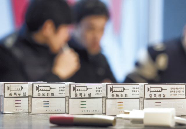 궐련형 전자담배는 담뱃잎을 그대로 쪄서 만든 것이므로, 독성이 약해질 수 없다고 전문가들은 주장한다. 궐련형 전자담배 사이로 궐련형 전자담배를 즐기는 흡연자들의 모습이 흐릿하게 보인다.  [연합뉴스]