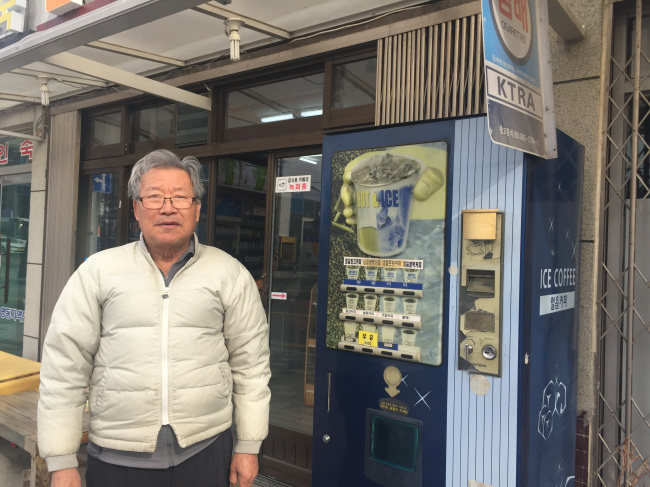 Choi Kyeong-hwan, who owns a convenience store named “Samho Super” near the Gangneung KTX station, poses with his coffee vending machine which is about 20 years old in front of his shop. (Claire Lee/ The Korea Herald)