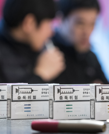 ‘아이코스’ 등 궐련형 전자담배는 담뱃잎을 그대로 쪄서 만든 것이므로, 독성이 약해질 수 없다고 전문가들은 주장해 왔다. 반면 담배업계는 궐련형 전자담배가 태우지 않아 일반 담배보다 90%가량 유해물질이 낮다고 홍보해 왔다. 최근 서울 시내 한 궐련형 전자담배 판매점. 제품들 사이로 궐련형 전자담배를 즐기는 흡연자들의 모습이 흐릿하게 보인다. [연합뉴스]