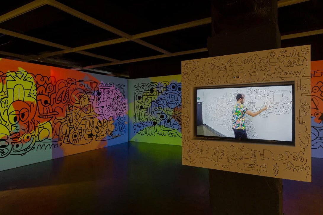 Inside of the “Fun Factory: Superstar Jon Burgerman” exhibition with a video playing to show the full process of the mural shown in the picture (https://www.jonburgerman.com/)