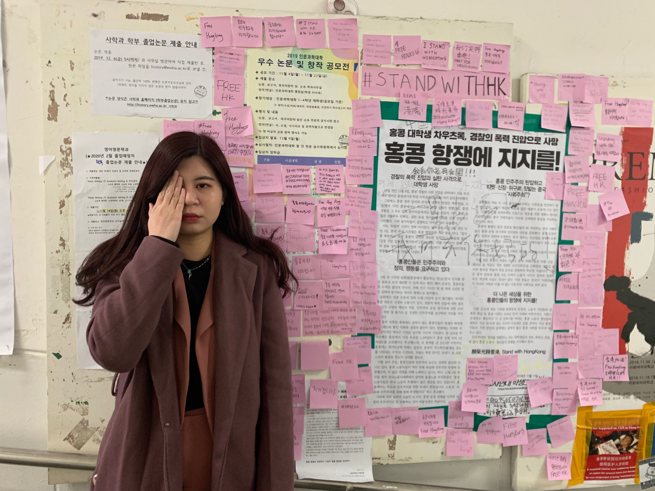 An international student from Hong Kong, poses in front of the Ewha Womans University wall covered with posters and sticky notes expressing support for Hong Kong protests. (Kim Arin/The Korea Herald)
