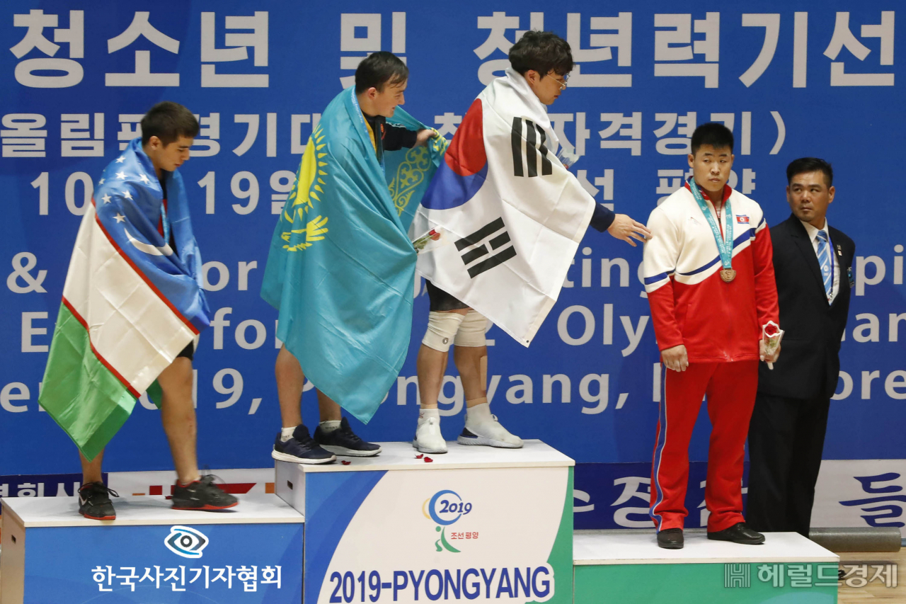 〈56회 한국보도사진상 - 최우수상〉 2019 아시아 유소년·주니어 역도선수권대회가 열리고 있는 25일 오후 평양 청춘가역도전용경기장에서 주니어 남자 89㎏급에 출전한 염다훈(한국체대)이 시상대에 올라 북측의 박금일 선수를 비롯한 수상자들에게 1등 시상대에 함께 서자고 권하고 있다. 염 선수는 이날 인상 160㎏, 용상 198㎏, 합계 258㎏을 들어올리며 인상 동메달, 용상과 합계 부문에 금메달을 추가했다. 이는 이번 대회 남측 선수단 최초 합계 금메달로 이날 처음 평양에 애국가를 울렸다. 〈이정아기자/ 한겨레/ 한국사진기자협회 - 무단전재 재배포금지〉