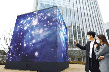 모델들이 전시장 외부 롯데월드타워 야외 공간에 실외용 LED 사이니지를 이어 붙여 만든 가로, 세로, 높이 각 6미터 크기의 미디어큐브를 통해 고(故)김환기 화백의 대표작 ‘우주’를 미디어아트로 구현한 작품을 감상하고 있는 모습. [LG전자 제공]