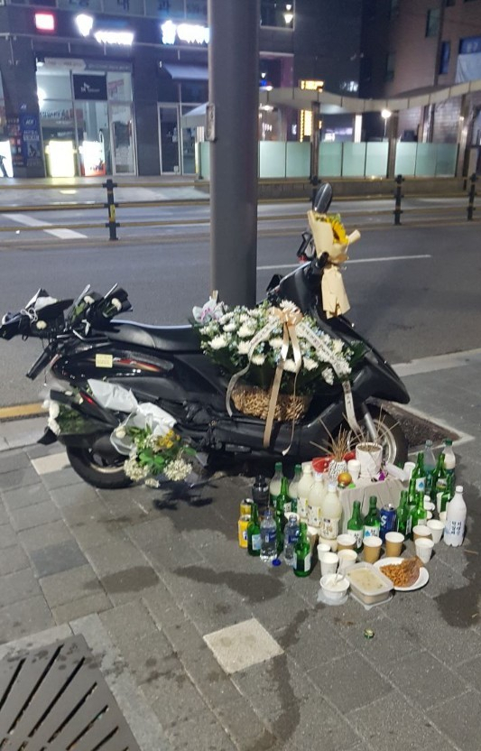 26일 한 배달라이더가 화물차에 치여 숨진 사고가 발생한 서울 강남구 지하철 선릉역 인근 모습. 같은 날 밤 숨진 라이더가 타던 오토바이 주위로 다른 라이더들이 추모의 의미로 꽃, 술 등을 올려놓았다. [민주노총 제공]