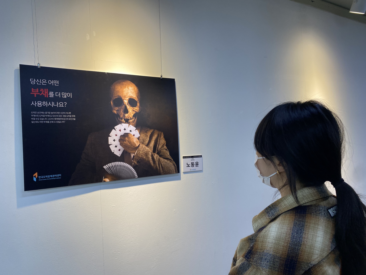 18일 을지로 아뜨리애 갤러리에서 관람객이 포스터 ‘당신의 부채’를 보고 있다. 본 포스터의 창작자 노동윤 작가는 포커 카드를 부채처럼 쓰는 도박중독자를 표현해 ‘2021년 도박문제 예방 공모전’의 청소년부 포스터 부문 장려상을 수상했다. [한국도박문제관리센터 제공]