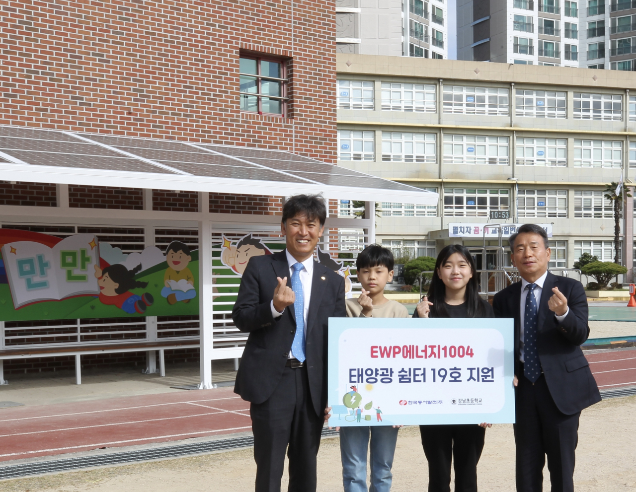 울산시 남구 강남초등학교에 설치된 'EWP에너지1004' 태양광 쉼터 19호 지원식에서 김영문 동서발전 사장(왼쪽에서 첫번째), 김욱년 강남초등학교장(왼쪽에서 네번째)과 강남초 학생들이 손가락하트 포즈를 취하며 기념사진을 촬영하는 모습. [동서발전 제공]