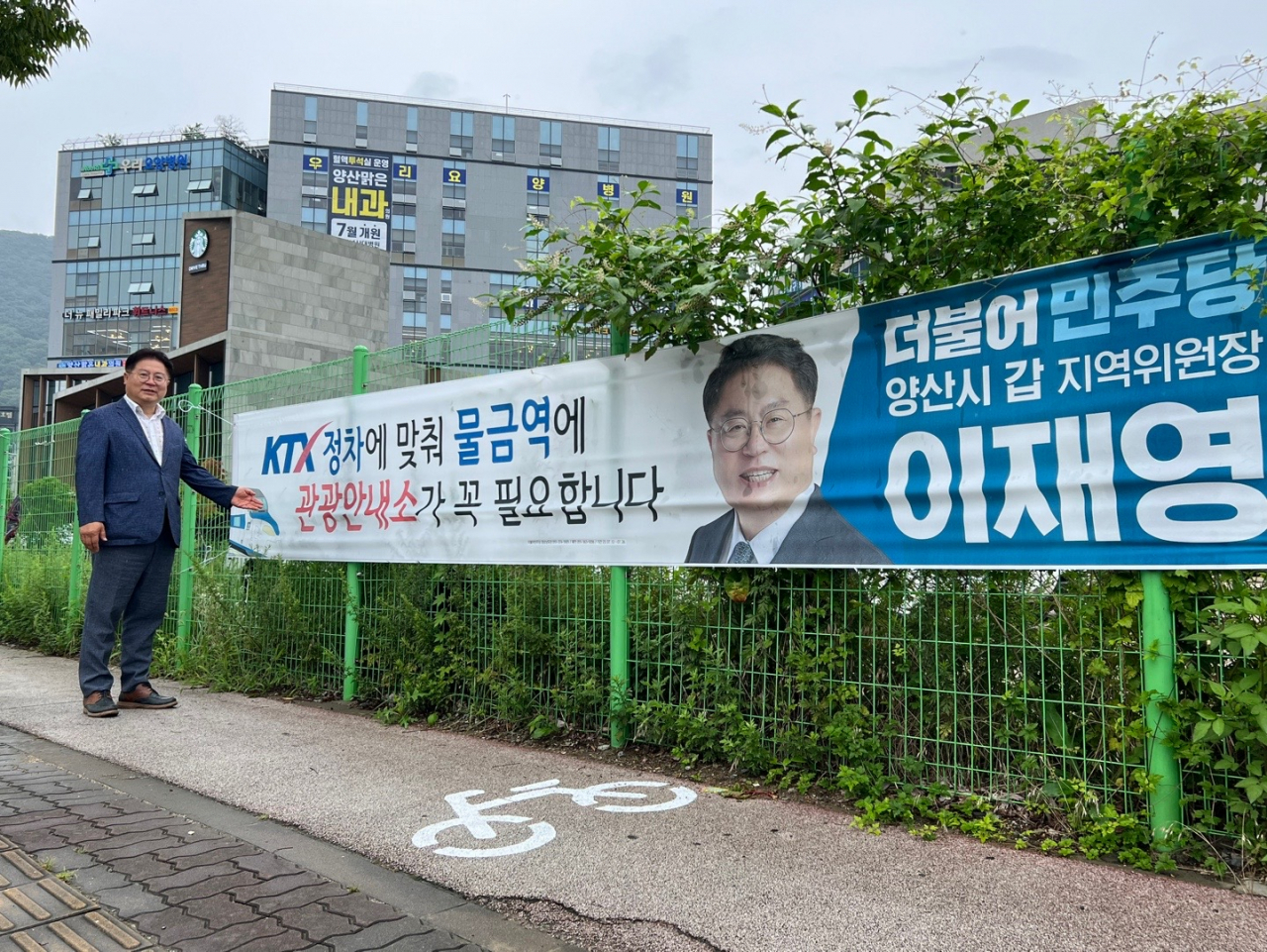 더불어민주당 양산 갑 지역 이재영 위원장이 KTX 정차예정인 물금역 시 관광안내소 설치를 주장하는 플래카드 앞에 서 있다. [사진=이재영 위원장]