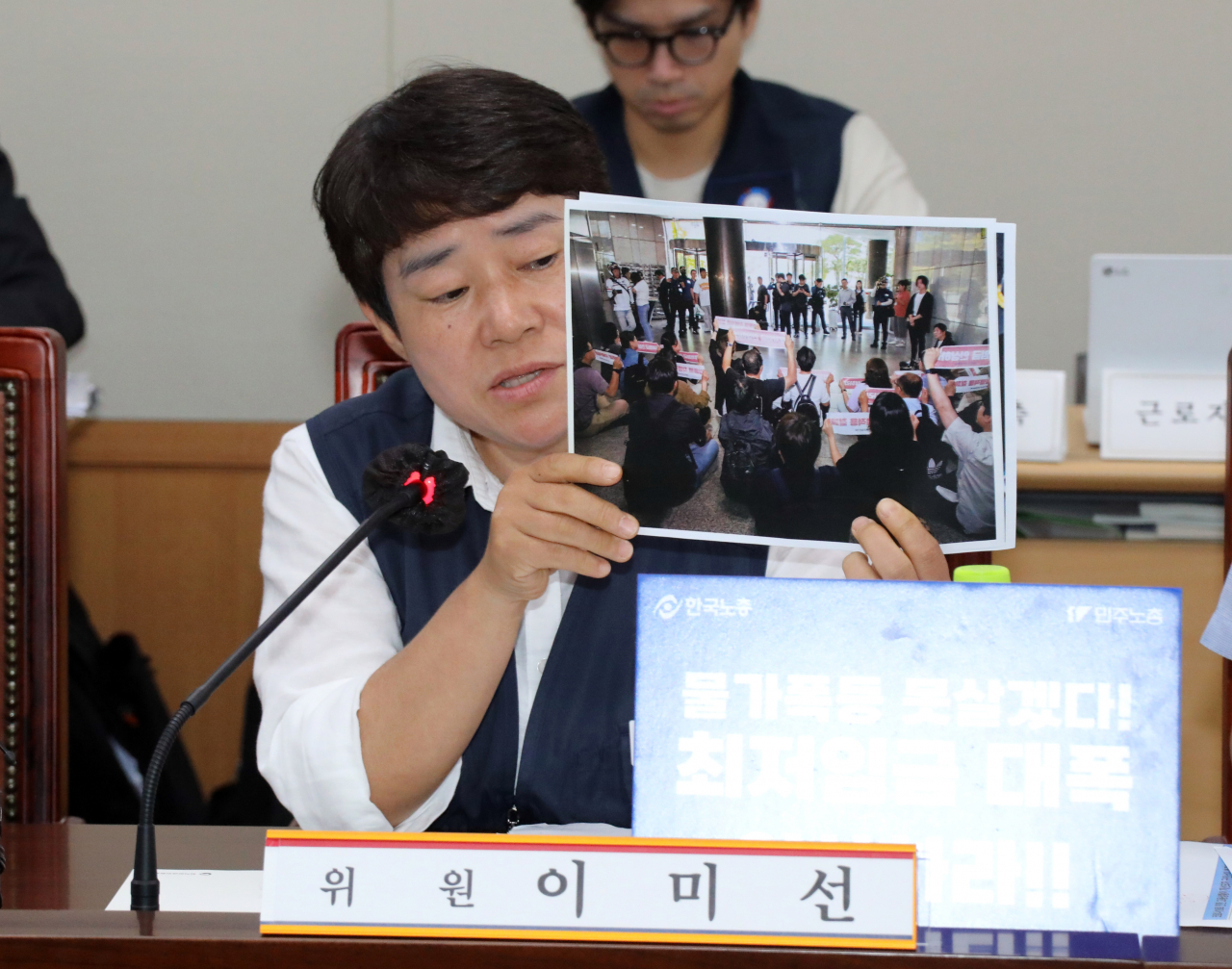 최저임금위원회 근로자위원 운영위원인 이미선 민주노총 부위원장(오른쪽)이 최저임금 법정 심의 기한인 27일 오후 정부세종청사 최저임금위원회에서 열린 제6차 전원회의에서 최저임금의 적용 확대와 인상을 요구하는 시위를 벌이다 끌려가는 장면이 담긴 사진을 보이며 발언하고 있다. [연합]