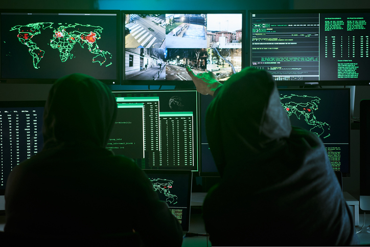 Back view of unrecognizable Criminal team Hacker pointing security cameras on screen monitor using computer for organizing massive data breach attack on government or big company servers. Dark room