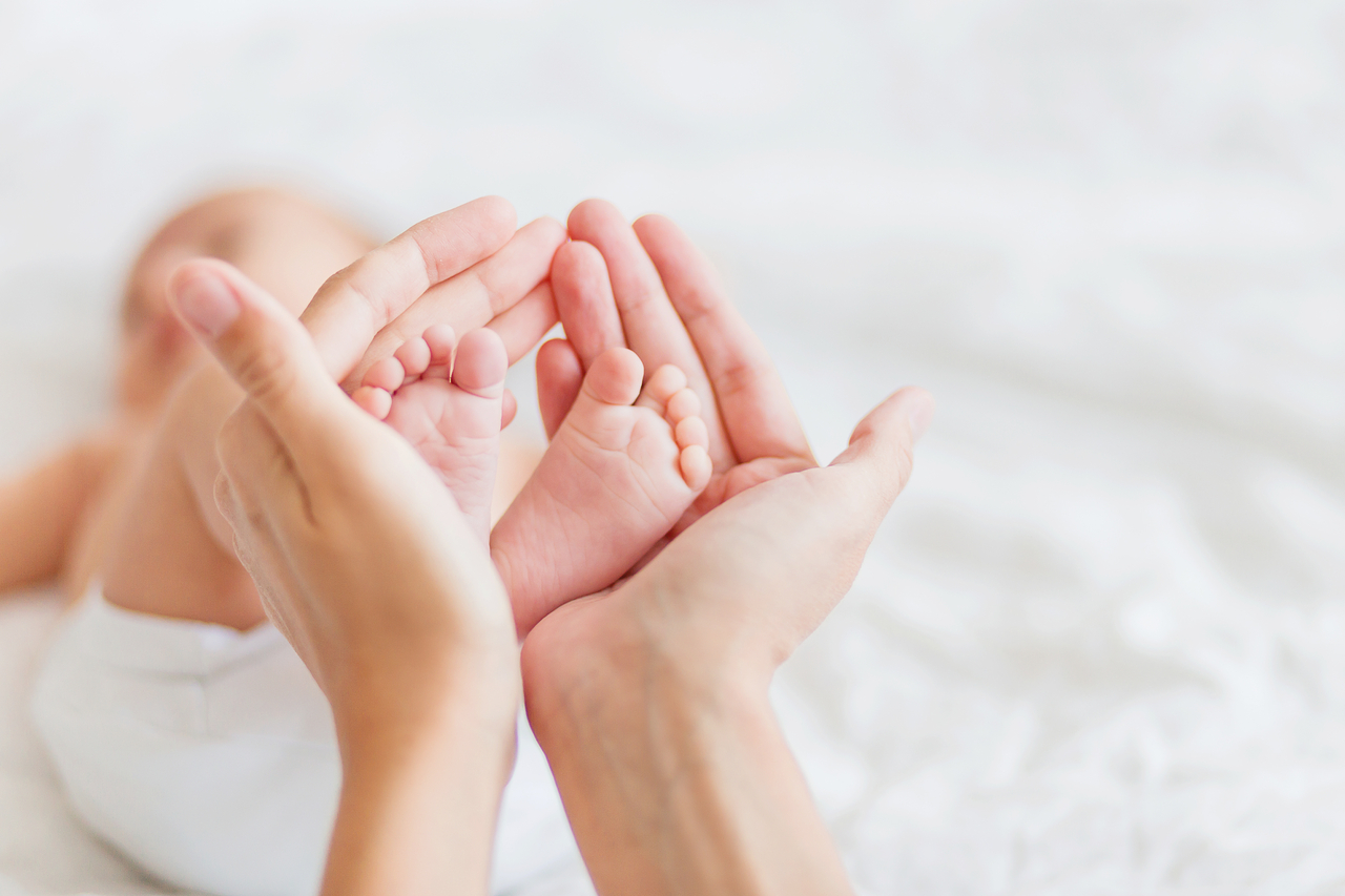 Mother holds newborn baby‘s bare feet. Tiny feet in woman’s hand. Cozy morning at home.