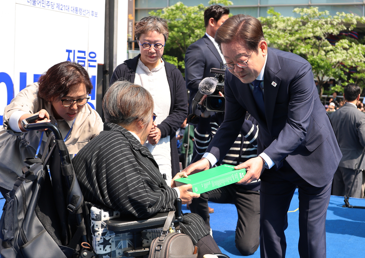 Democratic Party presidential candidate Lee Jae-myung receives a copy of "Green Book 2025" -- a policy proposal compiled by party think tank the Institute for Democracy -- from disability rights activist Park Kim Young-hee at a ceremony in Cheonggye Plaza, in Seoul's Gwanghwamun area Monday, the first official day of the presidential election campaign period for the June 3 election. (Yonhap)
