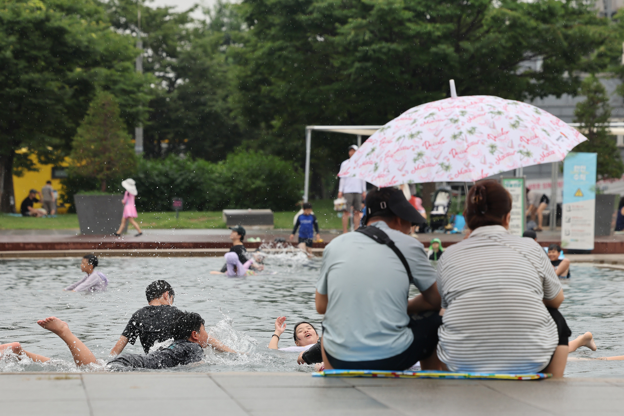 While two women sit by the pool under an umbrella at Yeouido Water Light Square in Seoul on Sunday, others continue to play in the water despite the rain. (Yonhap)