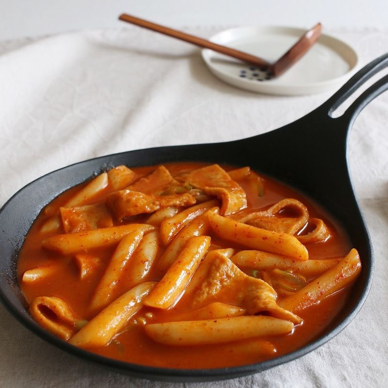 Classic Korean spicy tteokbokki with garaetteok cylinder rice cakes fish cakes and green onions simmering in bright red gochujang gochugaru sauce in a shallow pan with kelp broth as prepared in a Korea Herald recipe