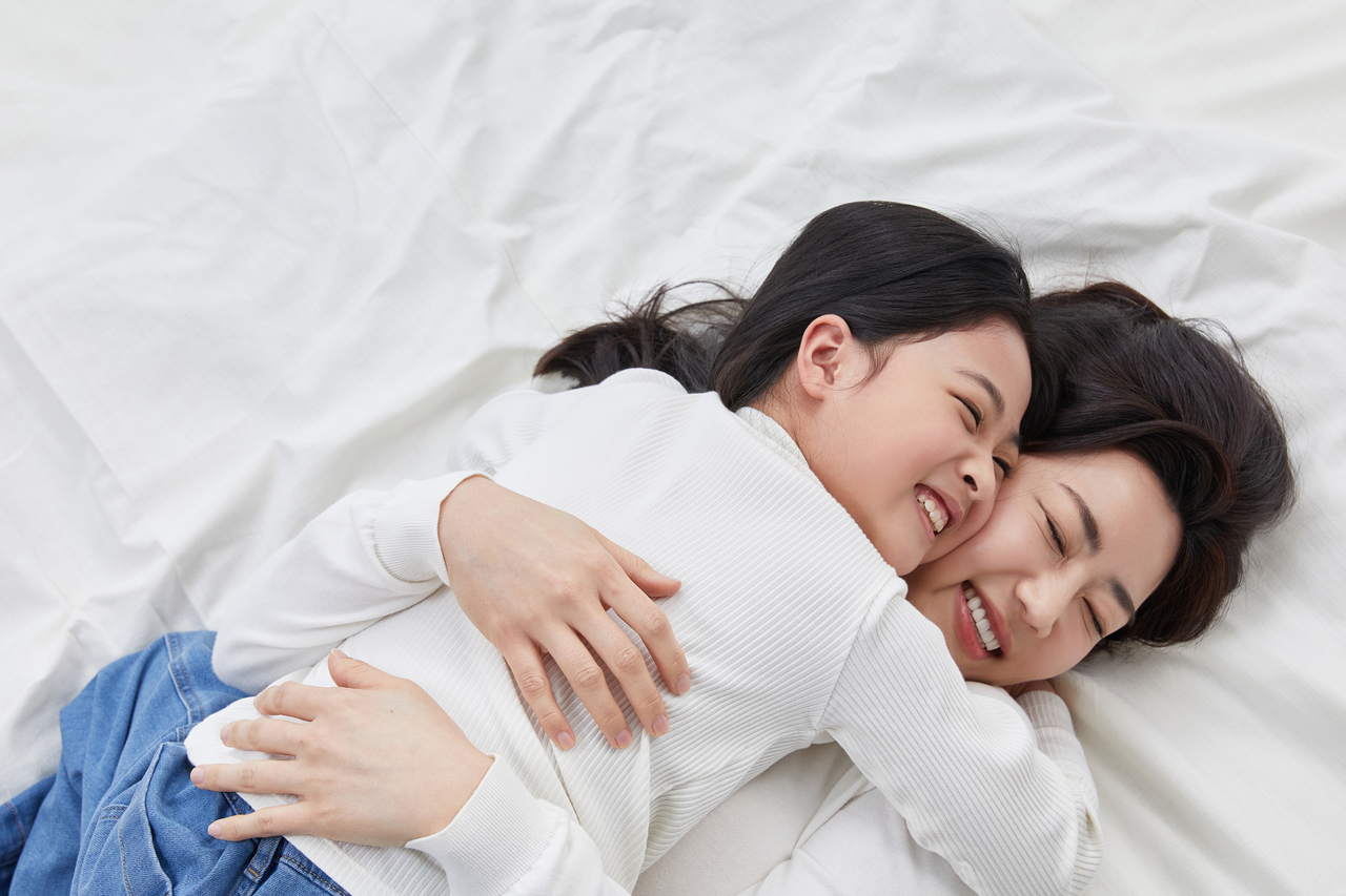 A mother and daughter lie on a bed, hugging and smiling. (123rf)