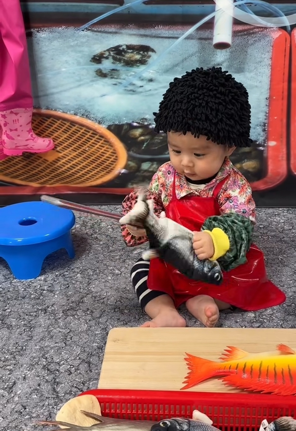 During a sensory play class, a baby girl in a wig, floral outfit and apron holds a toy knife and fish, resembling a fish vendor at a traditional market. (Instagram @from_g.u)