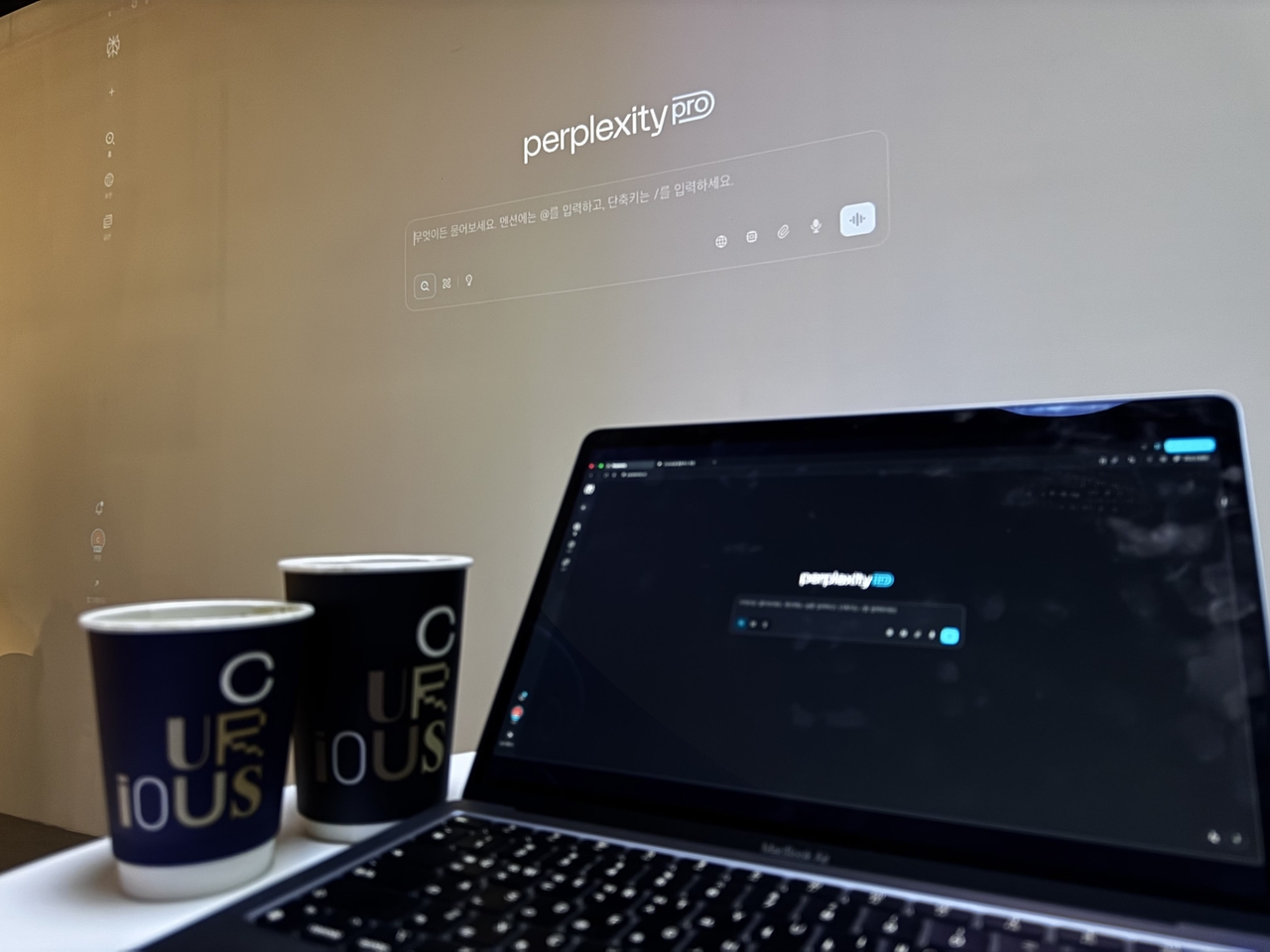 A laptop running Perplexity Pro sits in front of a wall-sized projection of the same interface, alongside coffee cups branded with the cafe’s name, Curious. (Moon Joon-hyun/The Korea Herald)