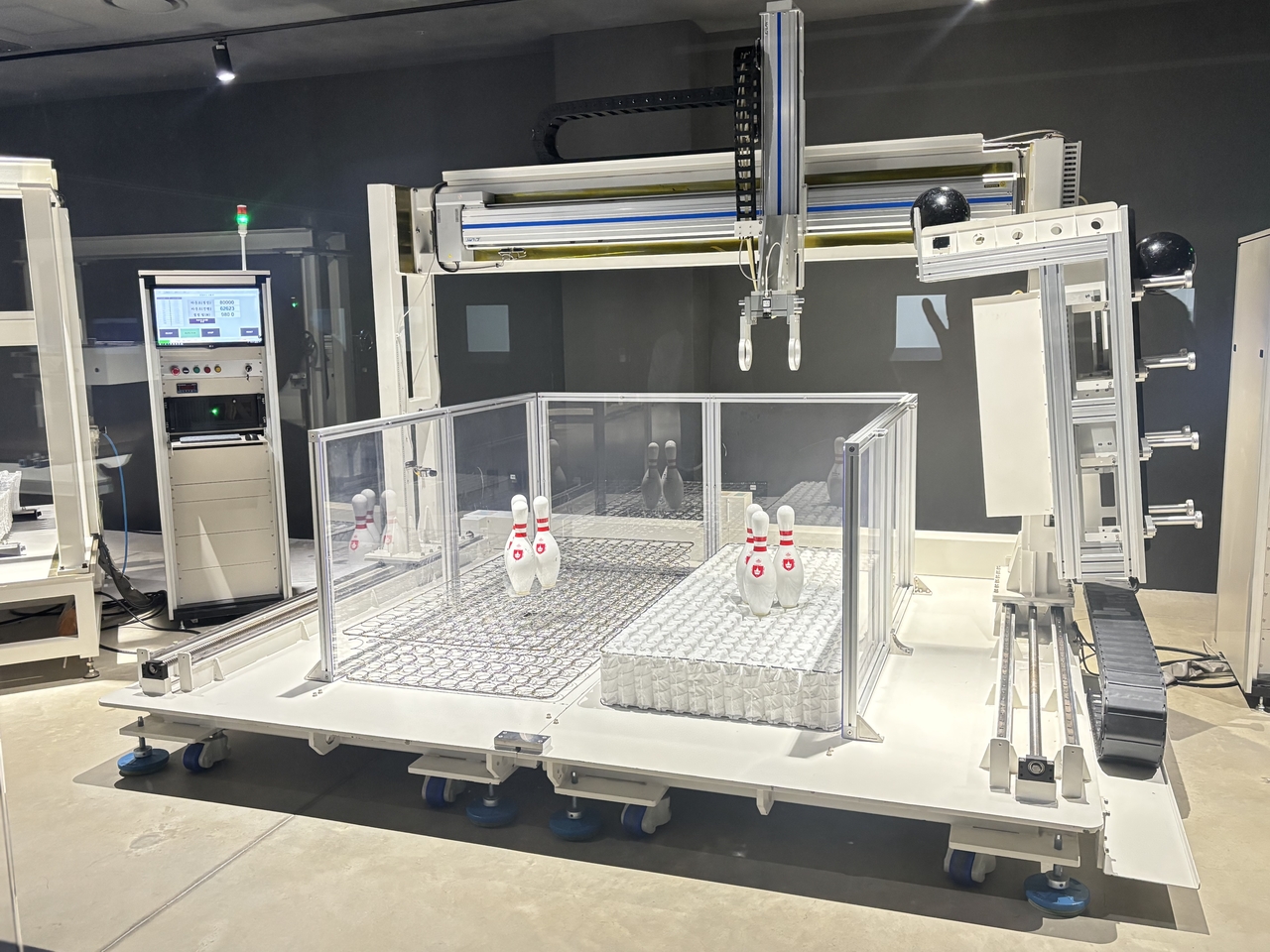 시몬스 팩토리움 내 R&D 센터에 설치된 실험 장치. 시몬스 침대 광고의 정점 볼링공 실험이다. 천장에서 내려온 로봇팔이 우측 선반에 놓인 볼링공을 1미터 높이에서 오른쪽과 왼쪽 매트리스 위에 각각 떨어뜨리고, 볼링핀이 계속 서있으면 성공 넘어지면 실패다. 오른쪽 매트(시몬스 제품)에 떨어진 볼링공은 볼링핀을 넘어뜨리지 못했지만, 왼쪽 매트 위에 떨어진 볼링공은 볼링핀 세개를 모두 넘어뜨렸다. [시몬스]