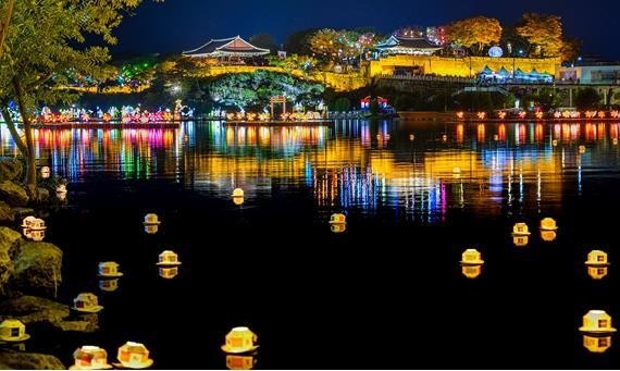 Massive illuminated lanterns depicting historical Korean figures floating on the Namgang River below Jinjuseong Fortress at night during the Jinju Namgang Yudeung Festival