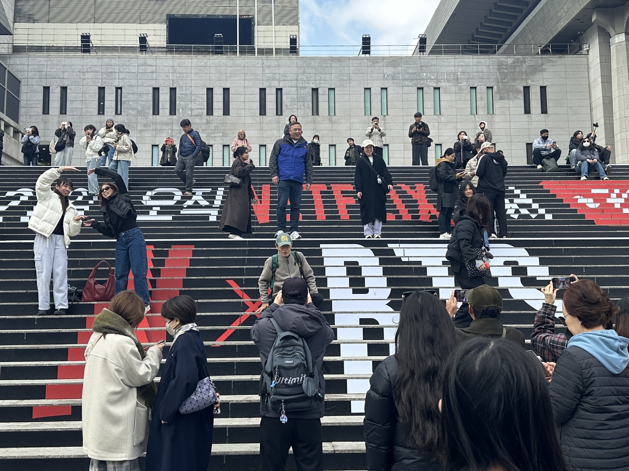Visitors pose for pictures on the stairs of Sejong Center for the Performing Arts, Friday, where promotional material for BTS' full-group comeback concert is displayed. (Lee Jung-joo/The Korea Herald)