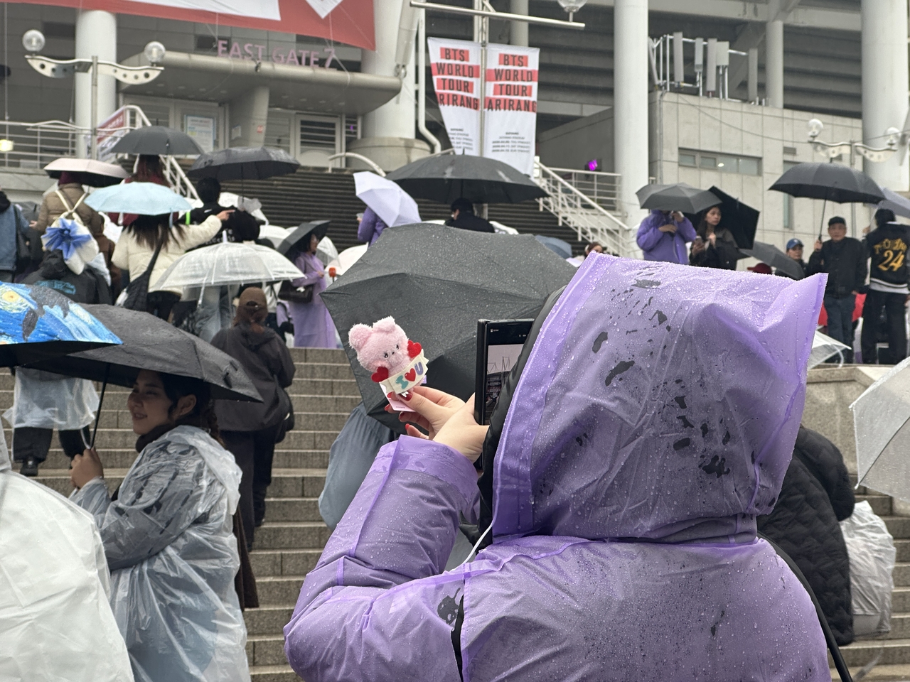 A BTS fan wearing a purple rain jacket takes a photo of a Cooky plushie, a character resembling member Jungkook, in front of Goyang Stadium, Thursday. (Lee Jung-joo/The Korea Herald)