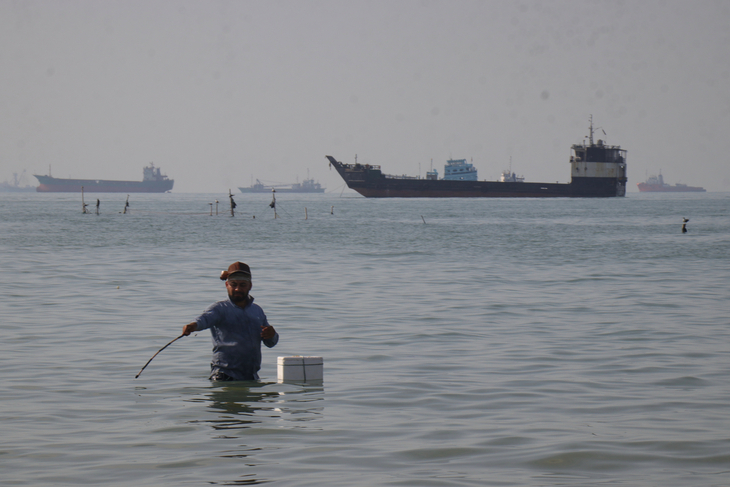 A man stands in the water, appearing to fish, as bulk carriers, cargo ships, and service vessels line the horizon in the Strait of Hormuz off Bandar Abbas, Iran, Monday, April 27. (AP-Yonhap)