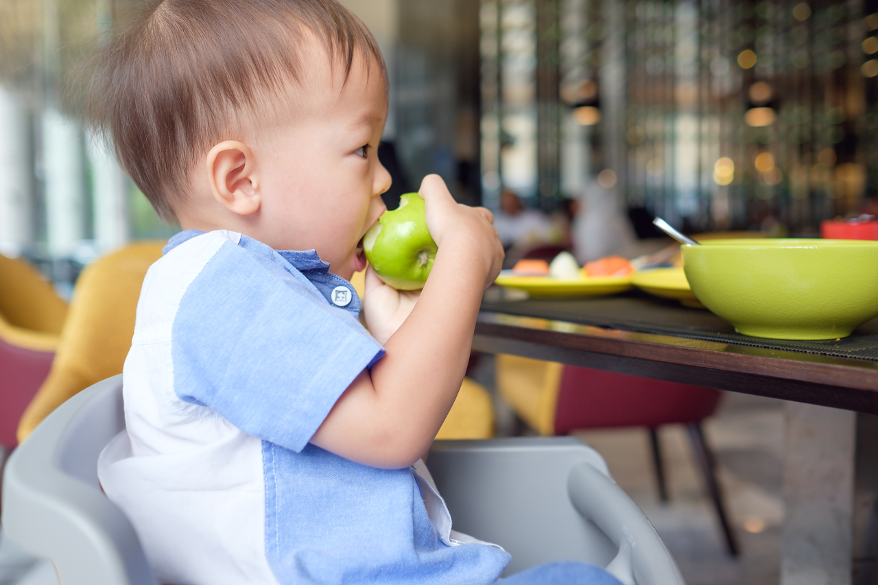 A toddler sits in a high chair eating a green apple. (Getty Images Bank)
