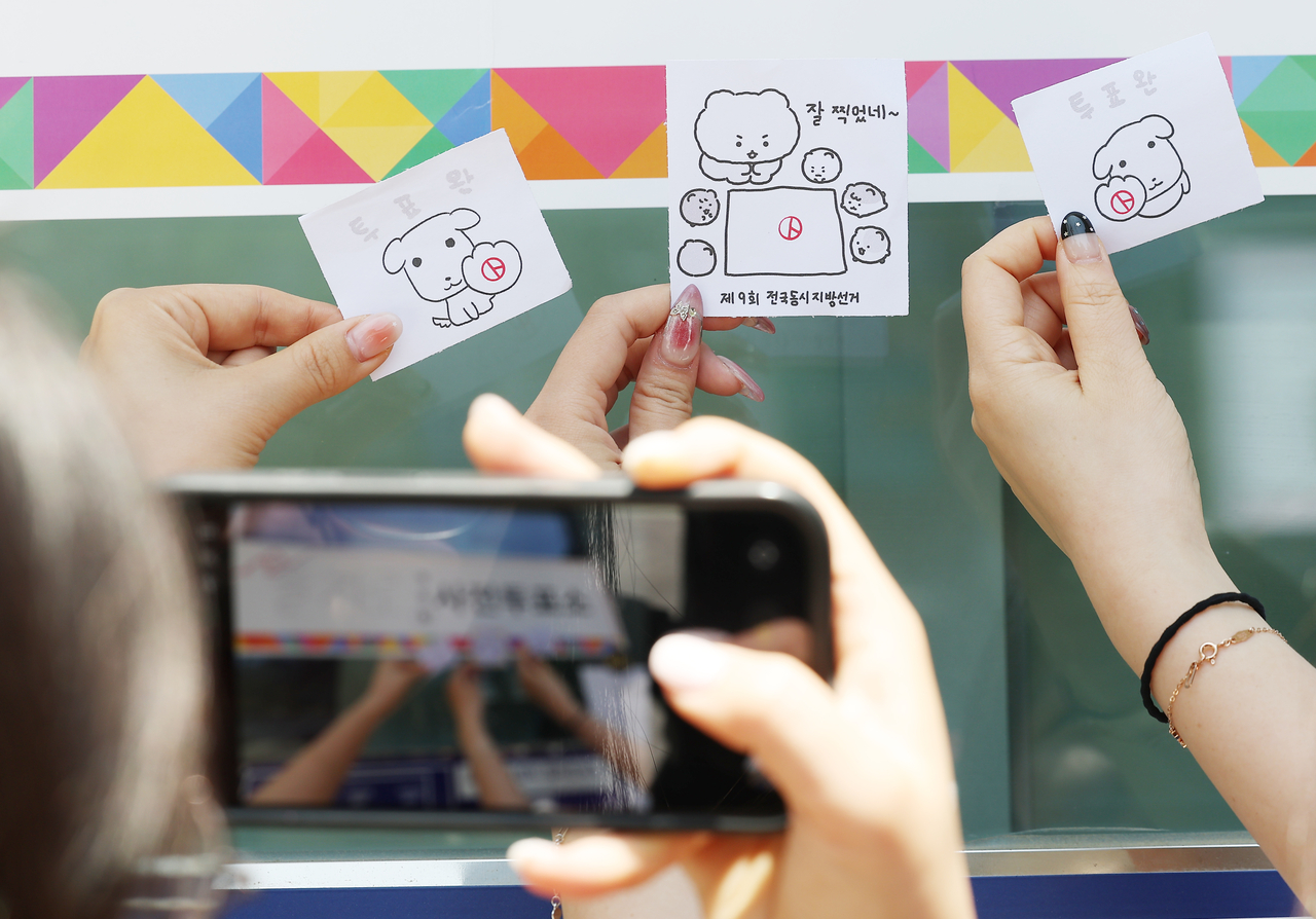 People take “voting proof” photos with character-themed cards stamped after voting outside a polling station in Seoul on Friday. (Yonhap)
