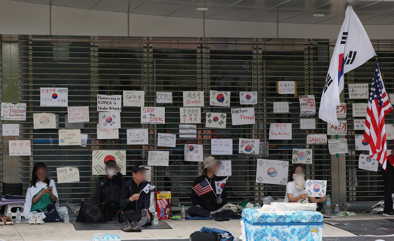 People rest in the shade by the closed gate of the Olympic Handball Gymnasium in Songpa-gu, Seoul, Wednesday, the sixth day of a protest blocking access to the site, which was used as a vote-counting center, as demonstrators called for a revote. (Yonhap)