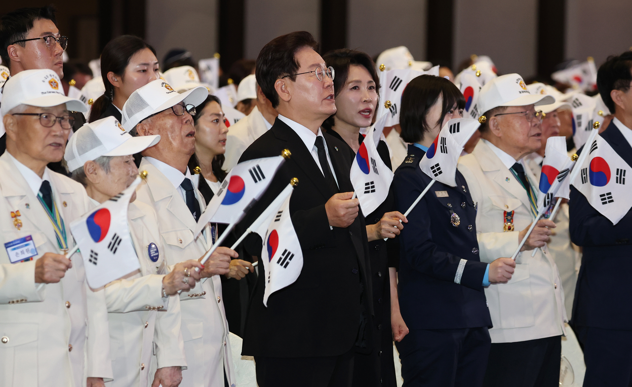 President Lee Jae Myung (center, in black suit) and first lady Kim Hea Kyung sing the commemorative "6.25 Song" while waving national flags alongside Korean War veterans and other attendees at a ceremony marking the 76th anniversary of the outbreak of the Korean War at the Suwon Convention Center in Gyeonggi Province, Thursday. (Yonhap)