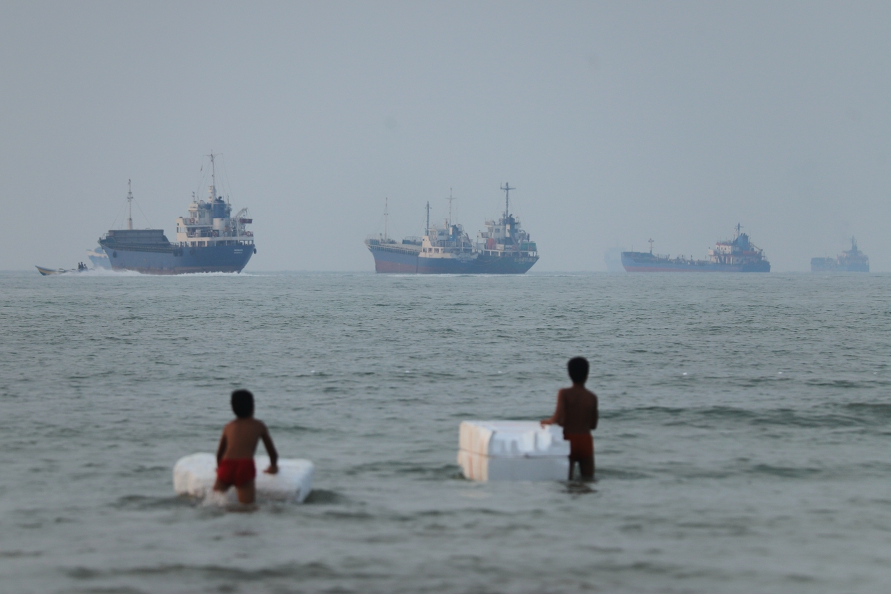 Two boys stand in shallow water with foam floats as cargo ships and other vessels are seen in the Strait of Hormuz off Bandar Abbas, Iran, Tuesday. (AP-Yonhap)