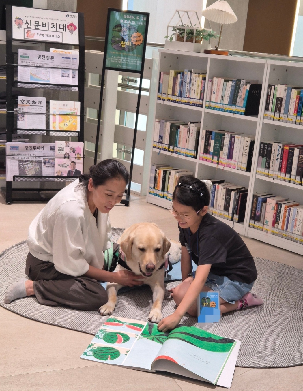 A young girl reads aloud to ChamE, a labrador retriever, as the dog's human companion, surnamed Shin, sits beside them during a Reading Education Assistance Dogs session hosted by the Korean Human-Animal Interaction Association at Achasan Forest Library in Seoul's Gwangjin-gu on June 30. (Choi Jae-hee/The Korea Herald)