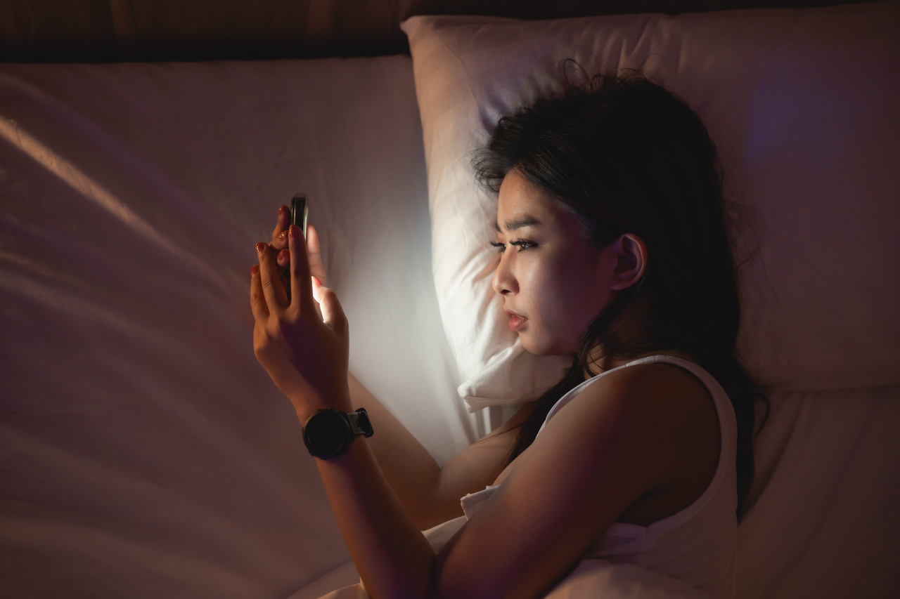 A woman looks at her smartphone while lying in bed. (123rf)