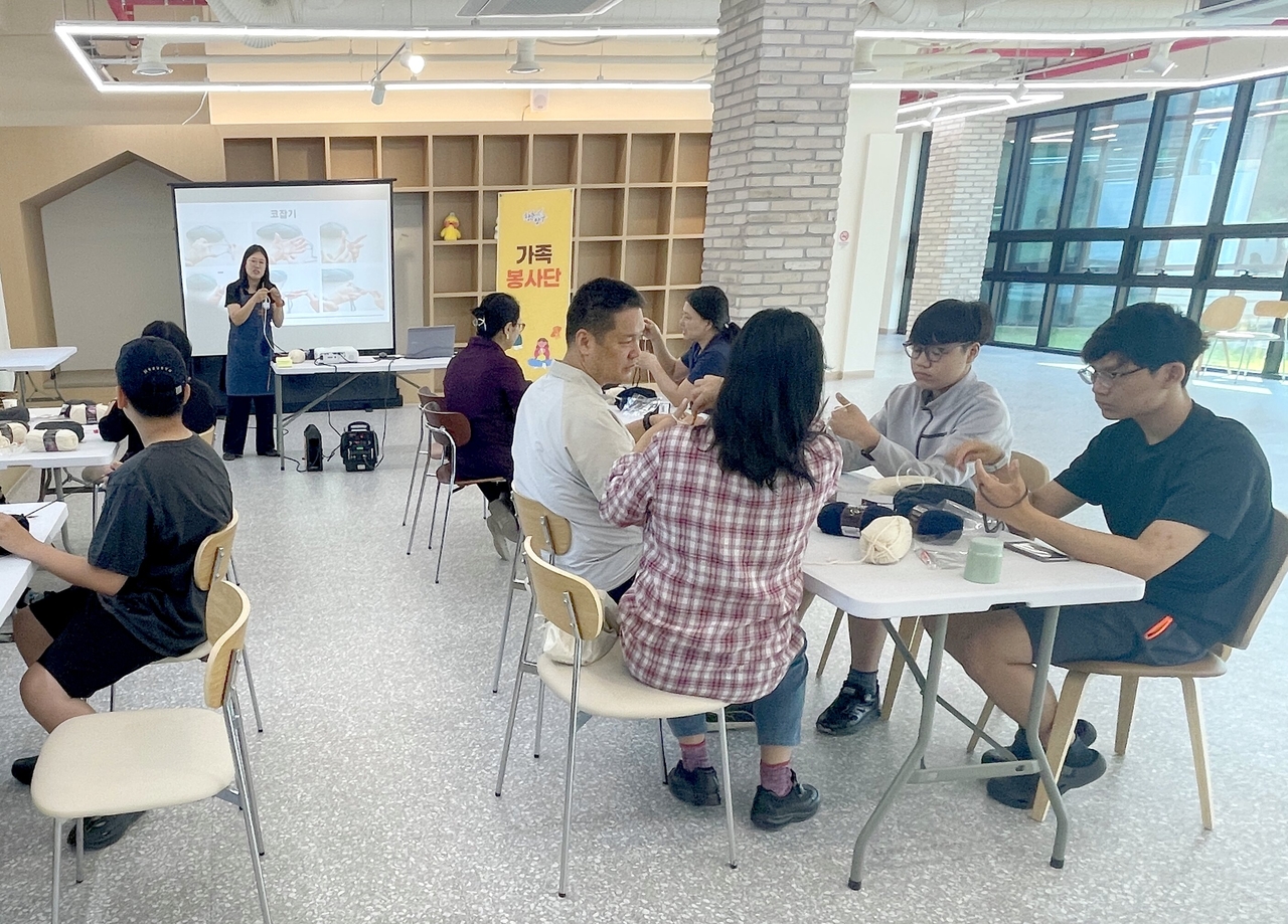 Family volunteers in Yanggu-gun knit with quiet concentration. The male participants wear expressions of unmistakable seriousness.