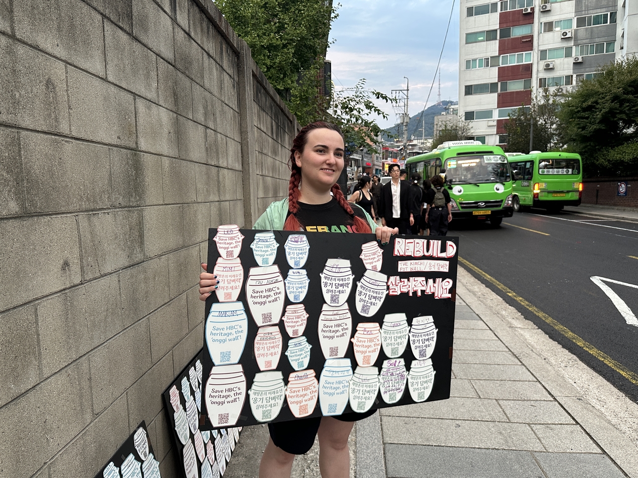 Sarah Mack holds a self-made poster board reading "Rebuild the kimchi pot wall" in Haebangchon, Yongsan-gu, Seoul, Sunday. People who submitted their views through the government’s online civil petition system added their names to paper onggi on the poster. (Tammy Park/The Korea Herald)