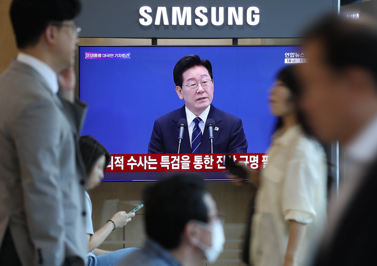 People walk by a screen showing President Lee Jae Myung's press conference at Seoul Station on Friday. (Yonhap)