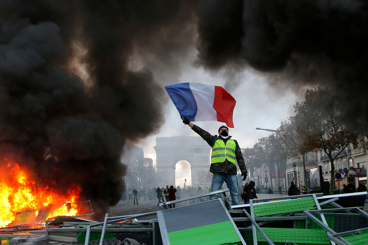 A protester in a yellow vest waves a French flag atop a burning barricade on the Champs-Élysées in Paris during a demonstration against soaring living costs. [AP]